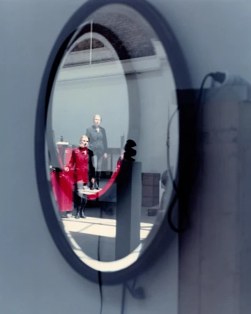 Reflection in a round mirror featuring two women in a room; one wears a red jumpsuit and the other a dark jacket.