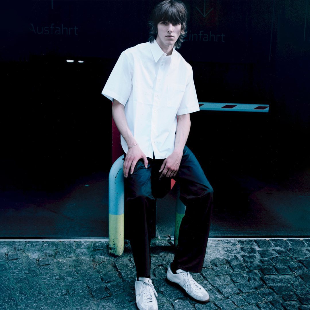A young man sits on a barrier wearing a white shirt, dark trousers, and white sneakers, posed in front of a dark wall with signage.