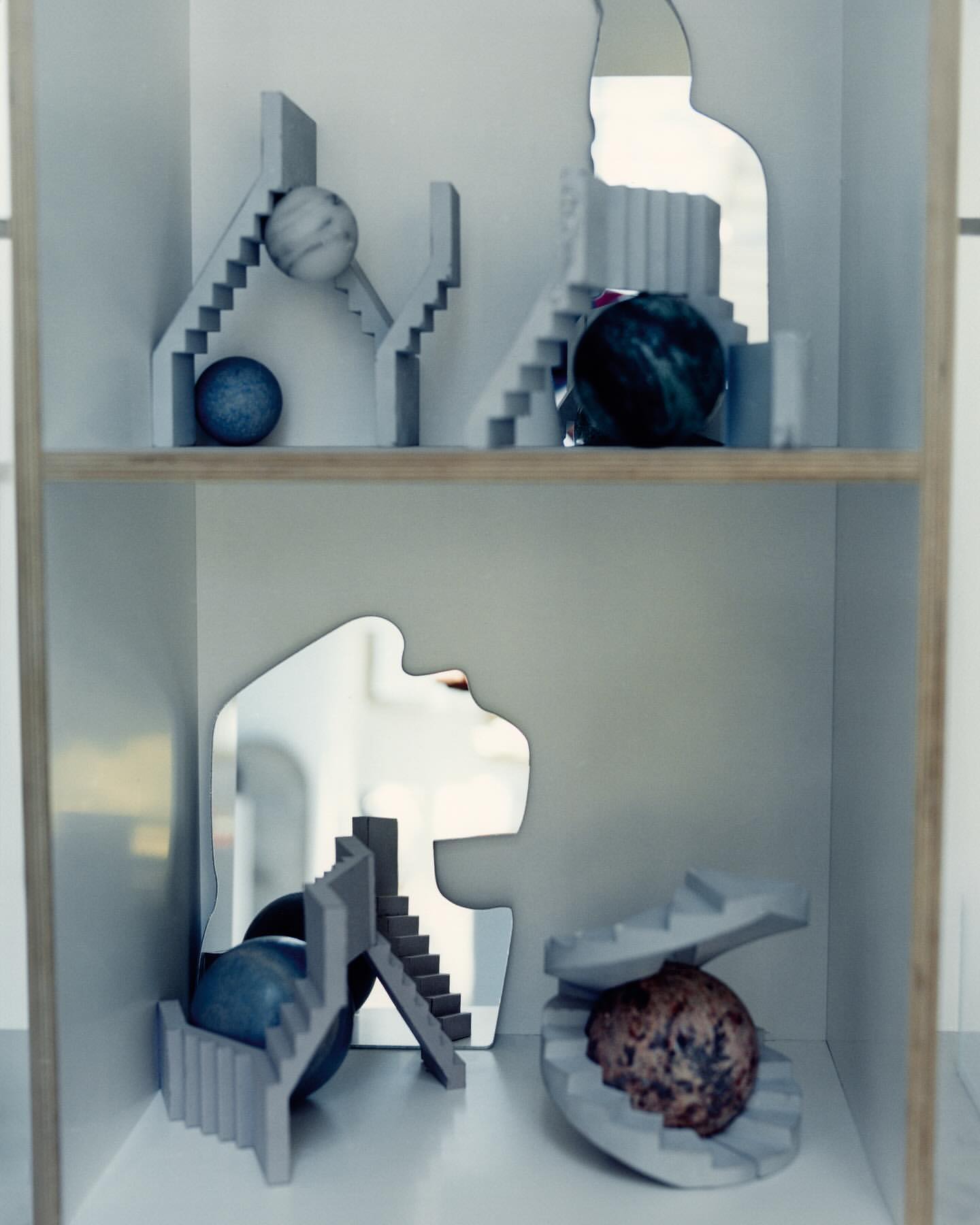 Still life of geometric objects on shelves, featuring spheres, staircases, and mirrors in a minimalist composition with muted colors.