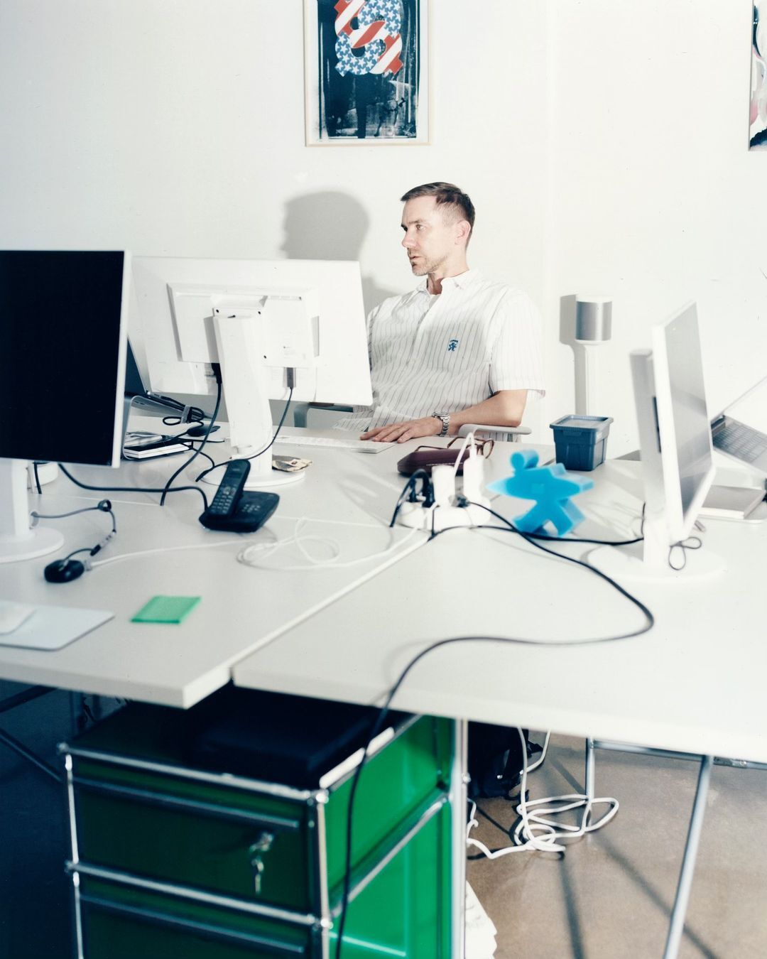 Man sits at a white desk with multiple computer monitors in an office space with white walls and a colorful poster in the background.