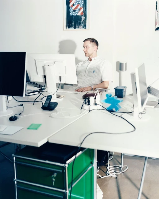 Man sits at a white desk with multiple computer monitors in an office space with white walls and a colorful poster in the background.
