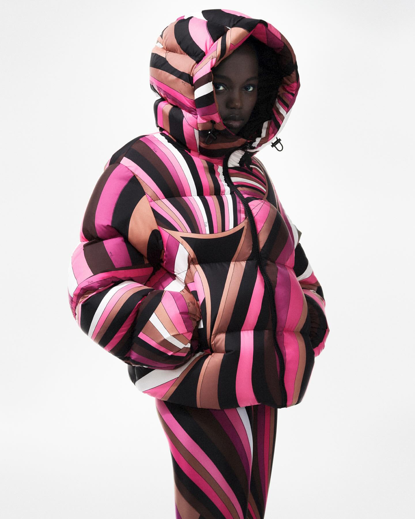 Fashion shot of a black female model wearing a pink and brown swirled puffer jacket and pants in a studio setting against a white backdrop.