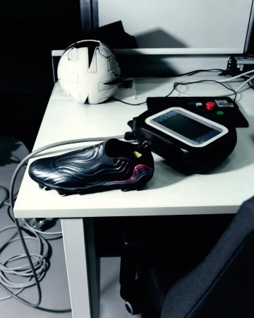 A dark cleat-style soccer shoe sits on a white table next to a black device and a soccer ball, showcasing a sports product.