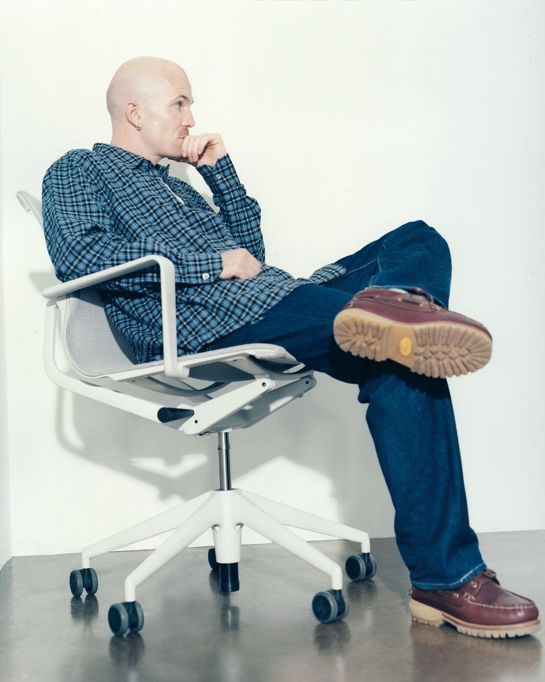 Bald adult male in a blue-and-grey flannel shirt sitting cross-legged in a white rolling office chair in a bright indoor setting.