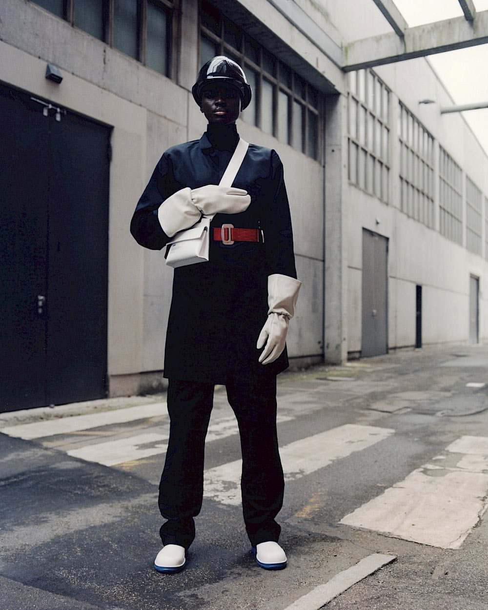 A person wearing a black outfit with a white bag and white shoes stands in an urban area, with a gray building in the background.