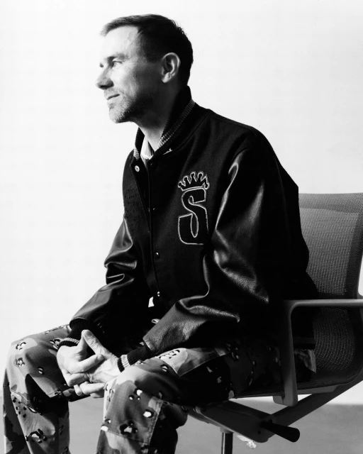 Monochrome portrait of an adult male seated wearing a letterman jacket and patterned pants against a simple studio backdrop.