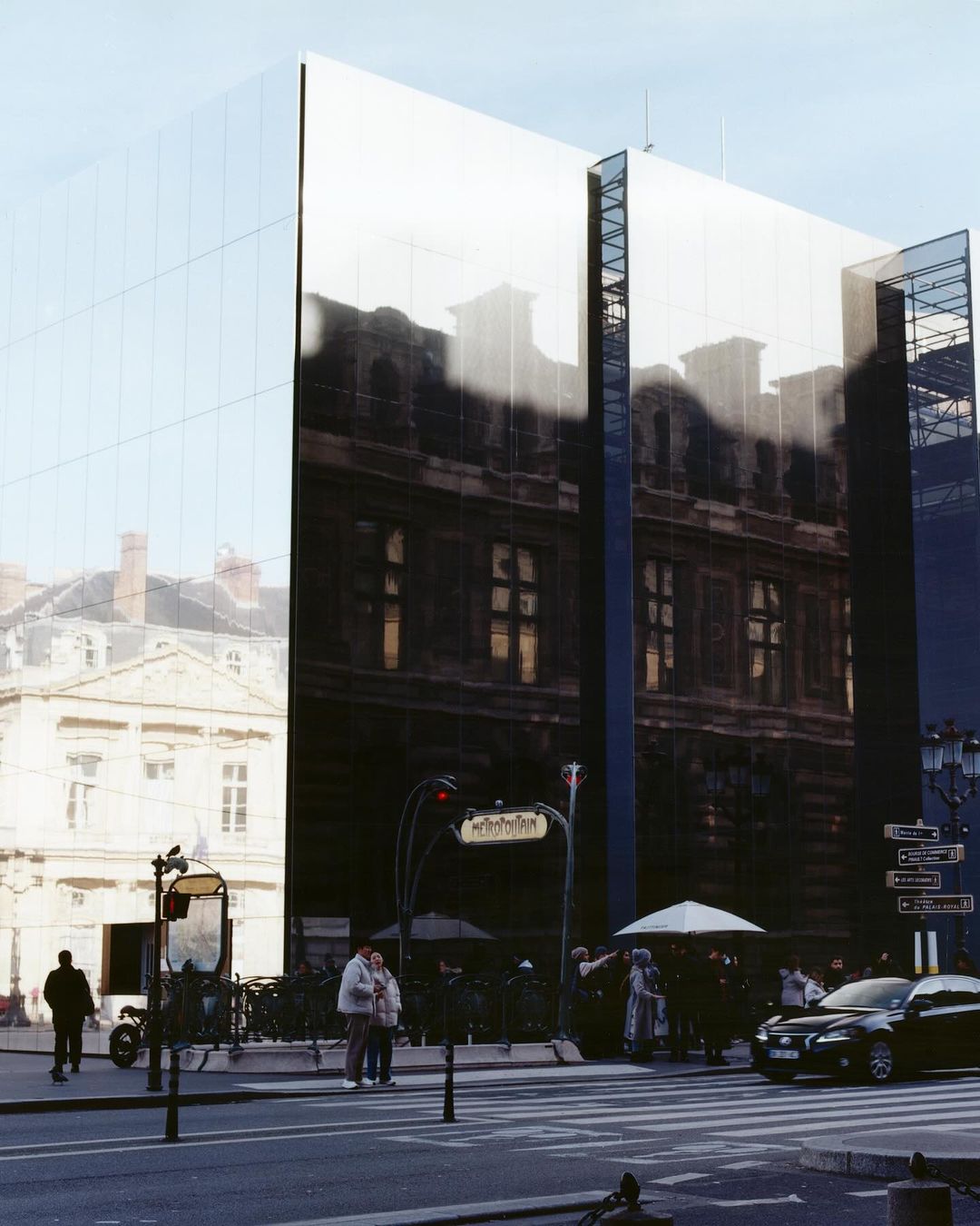 A modern mirrored building reflects the surrounding cityscape in Paris. Pedestrians and a metro station are visible on the street.
