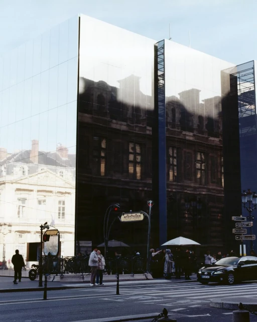 A modern mirrored building reflects the surrounding cityscape in Paris. Pedestrians and a metro station are visible on the street.