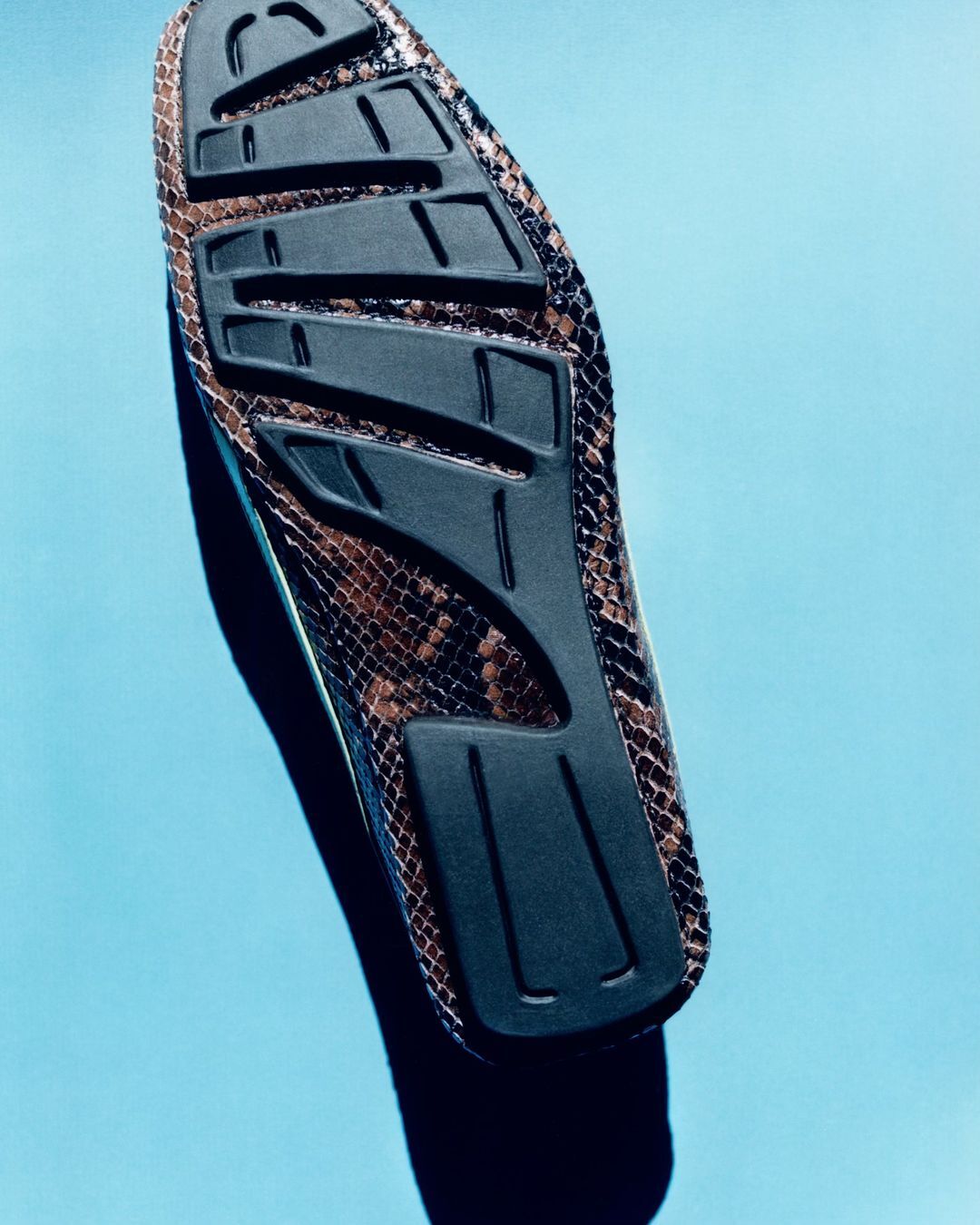 Brown snakeskin-patterned slip-on shoe with a black sole, displayed at an upward angle against a light blue backdrop in a studio setting.