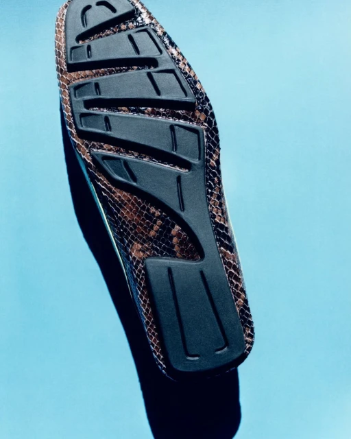 Brown snakeskin-patterned slip-on shoe with a black sole, displayed at an upward angle against a light blue backdrop in a studio setting.