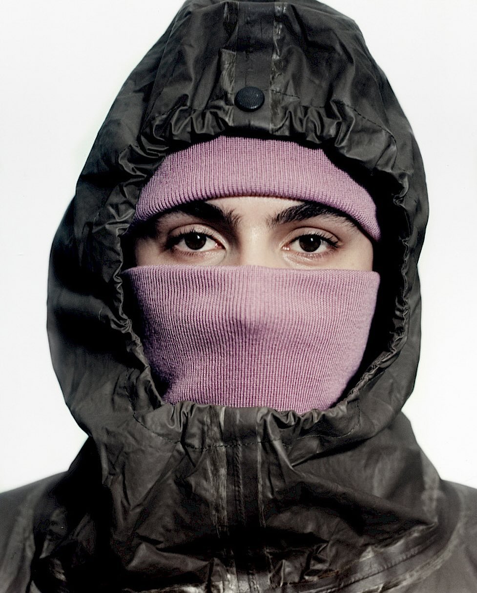 Close-up shot of an adult with dark eyes wearing a pink balaclava and a dark hooded jacket against a plain white background.