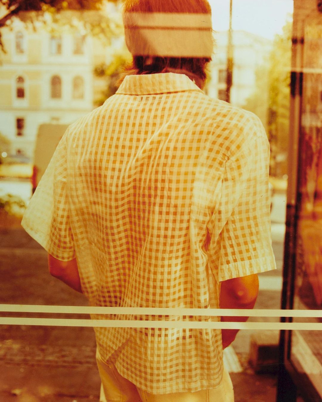 Back view of a person in a checkered shirt standing indoors with a building and trees seen through the window, filtered with a warm tone.