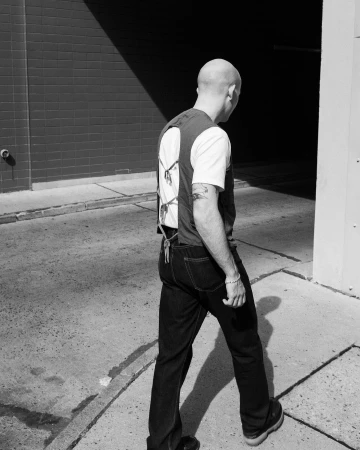 Bald man walking outdoors wearing dark jeans and a sleeveless top over a white t shirt, photographed by Maximilian Semlinger.