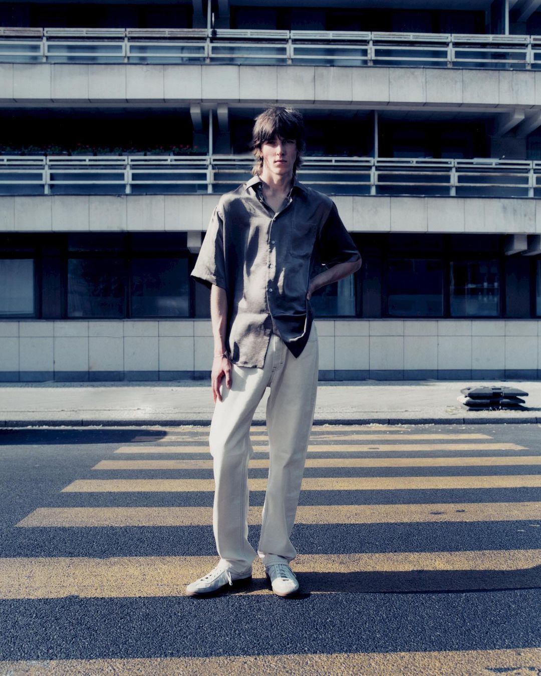Male model stands on crosswalk in casual button shirt and white pants in daylight urban fashion shoot with concrete building backdrop.