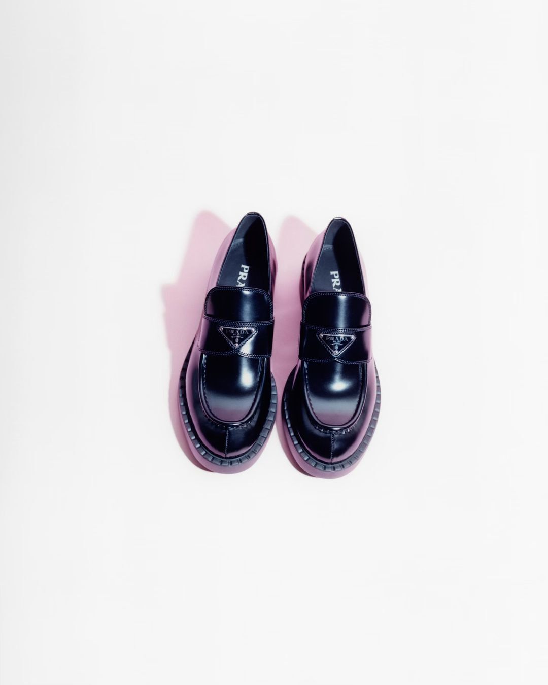 Two black Prada loafers with the brand's logo displayed in the center of each shoe, arranged on a light pink backdrop.