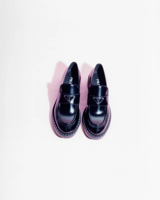 Two black Prada loafers with the brand's logo displayed in the center of each shoe, arranged on a light pink backdrop.