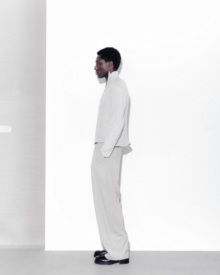 Fashion shot of a Black male model in a light grey jacket and pants posing in a minimalist photo studio with white walls and soft lighting.