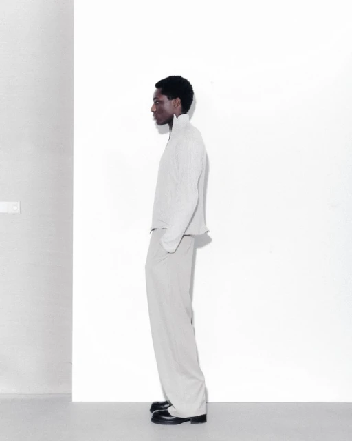Fashion shot of a Black male model in a light grey jacket and pants posing in a minimalist photo studio with white walls and soft lighting.