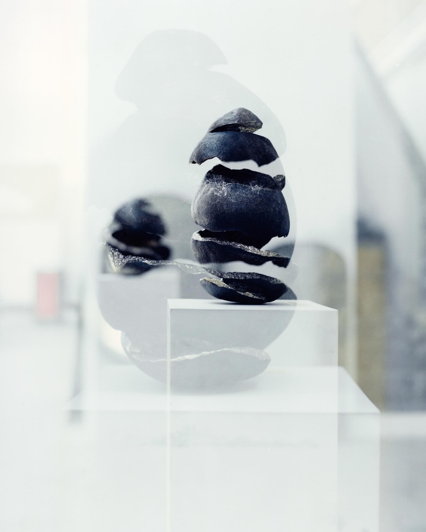 Abstract still life of stacked dark stones on white pedestals with a muted indoor background, creating a serene and minimalist aesthetic.
