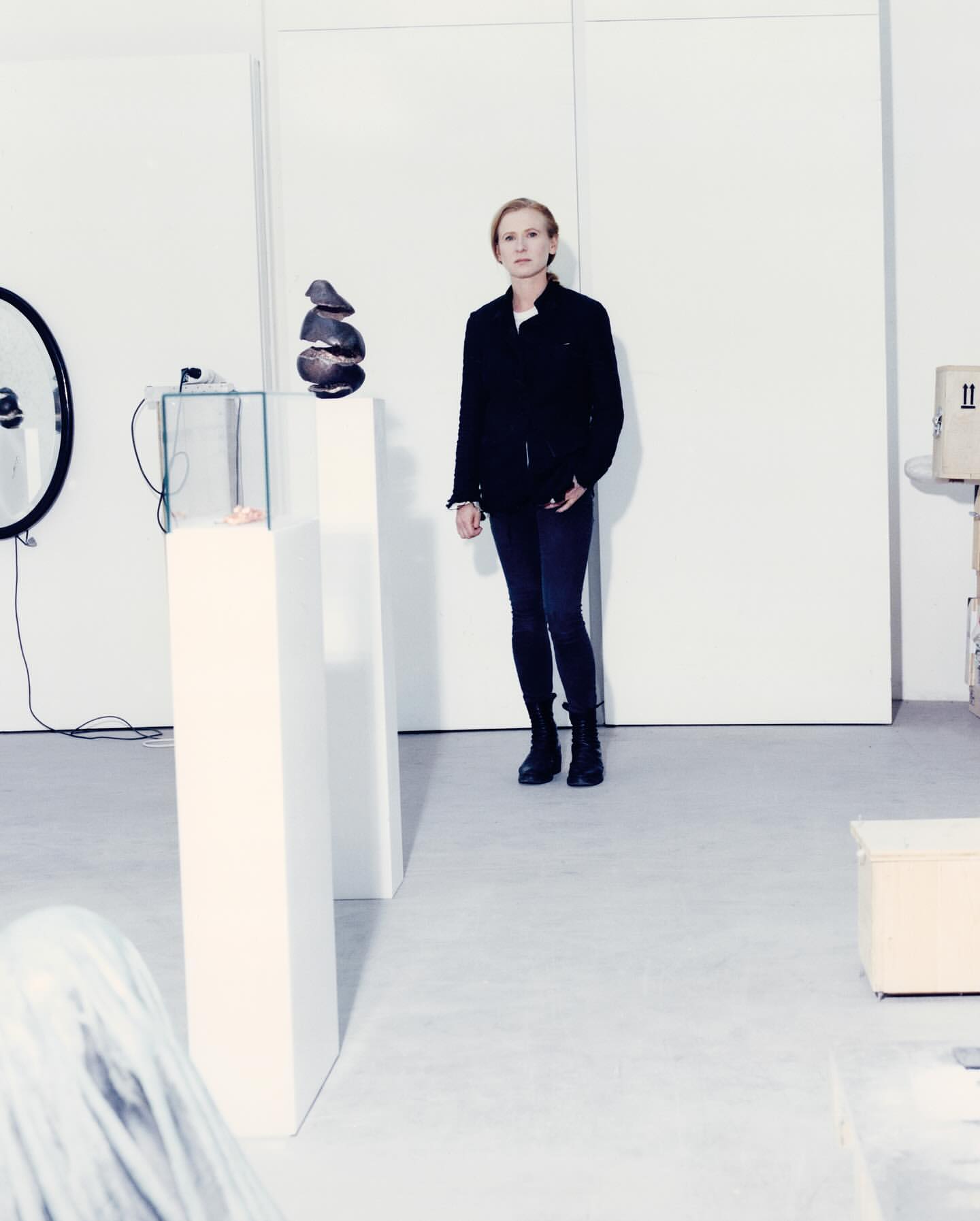 Woman in black jacket stands in a white art gallery with sculptures on pedestals, minimalist modern setting, and neutral color palette.