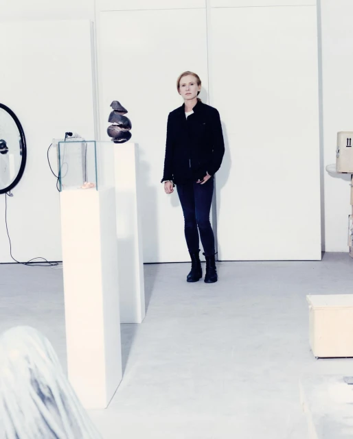 Woman in black jacket stands in a white art gallery with sculptures on pedestals, minimalist modern setting, and neutral color palette.