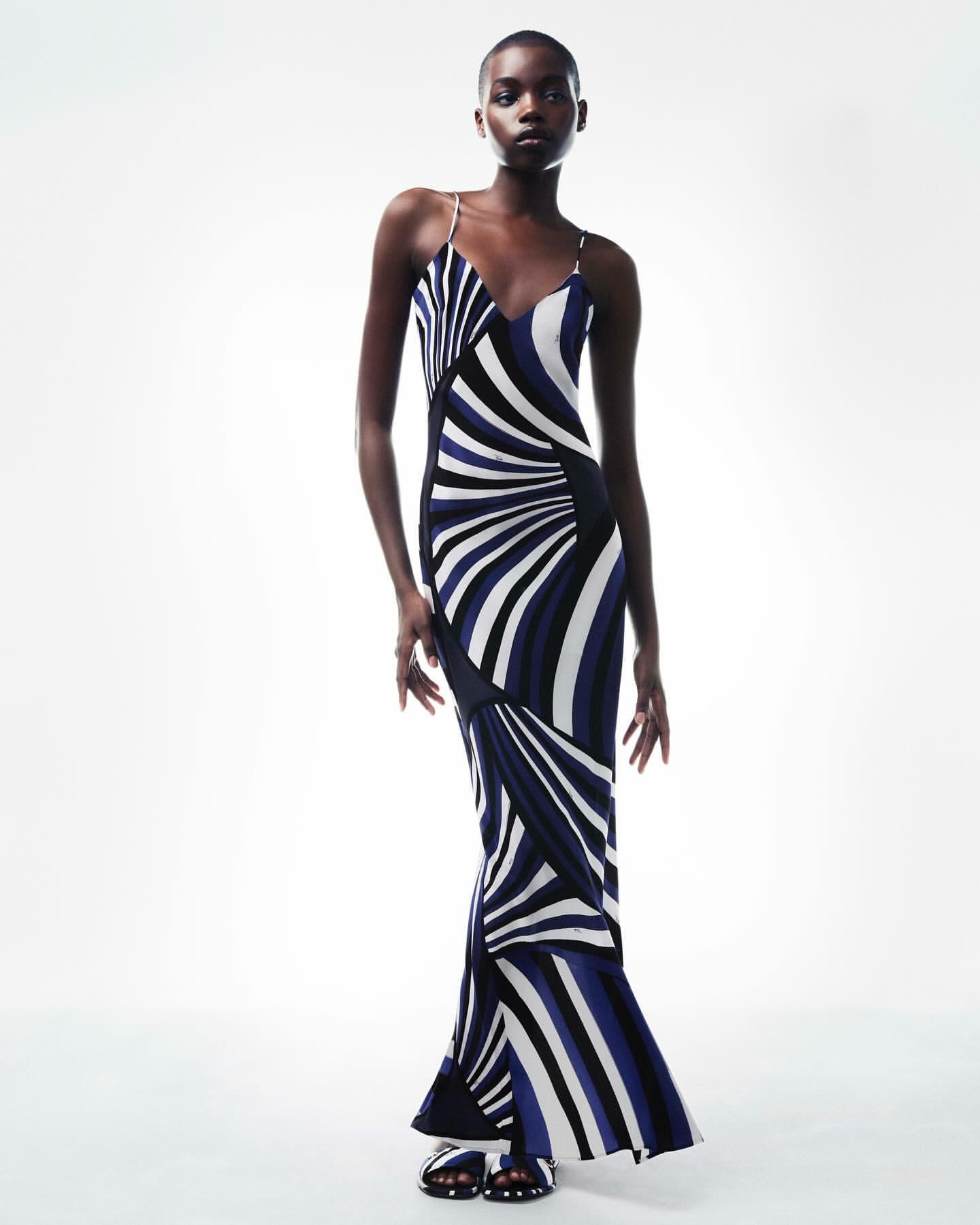 Elegant adult woman in a modern, striped maxi dress poses in a bright photo studio. Fashion editorial image with a minimalist background.