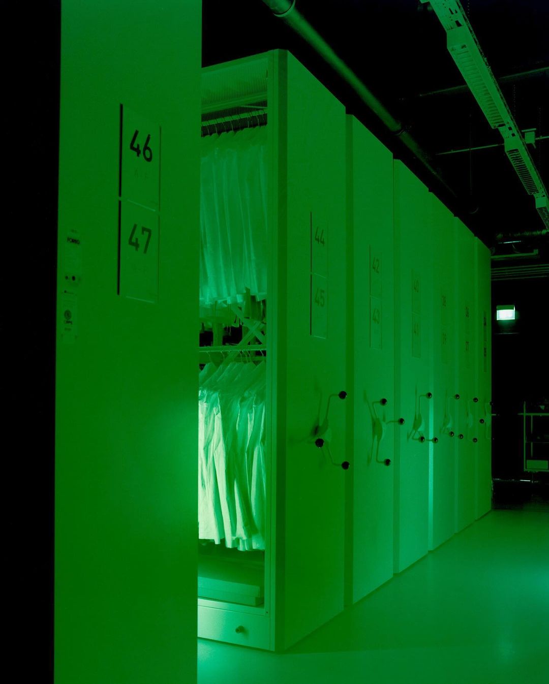 Long row of white storage cabinets in a dark room with a green light. Racks of white shirts hang inside one open cabinet door.