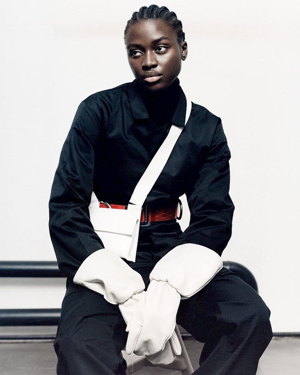 A solitary, dark-skinned model in a sleek black top and pants is seated in front of a white wall, accessorized with a white purse and gloves.