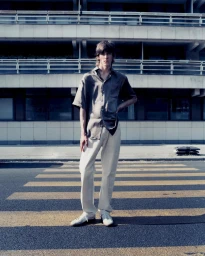 Young man in dark satin short-sleeve shirt and white trousers standing on a paved road by a building.