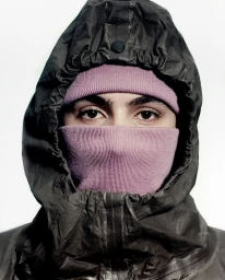 Close-up portrait of a person wearing a pink balaclava under a dark hooded jacket against a white background