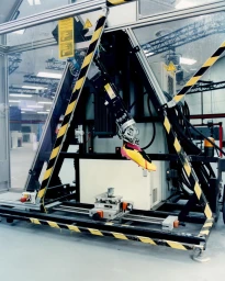 Robotic testing arm holding a yellow sports shoe inside an industrial testing rig with black and yellow hazard tape.