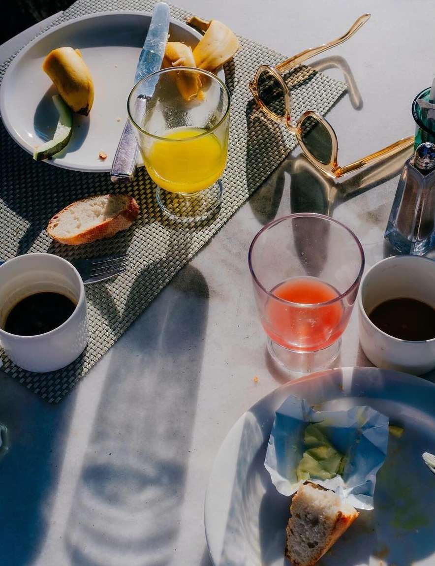 Overhead shot of breakfast table with plates, glasses of juice, coffee cups, bananas, bread, silverware, sunglasses, and salt shaker.