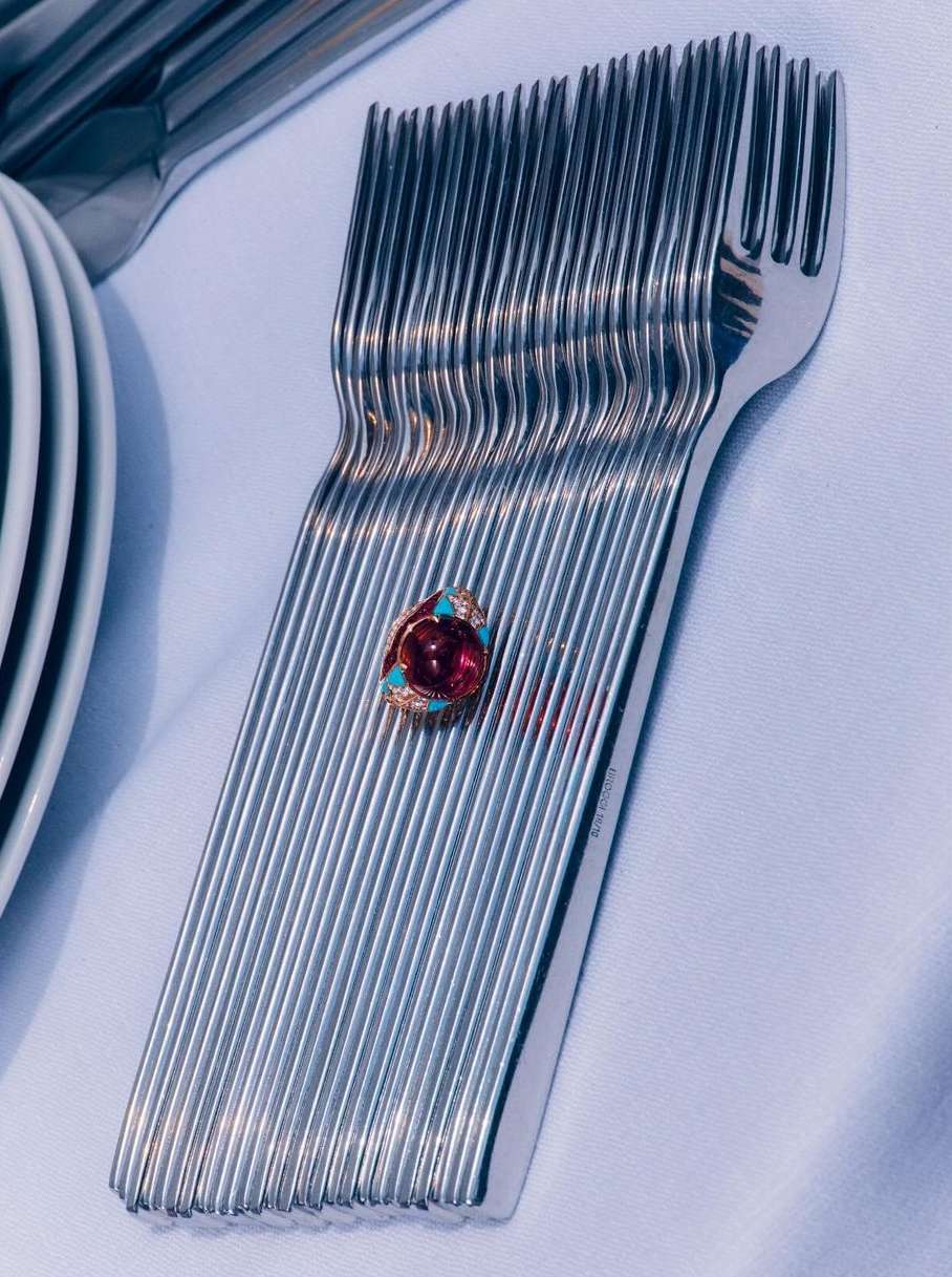 Close-up of silver forks stacked together, topped with a red and blue gemstone ring on a white tablecloth surface.