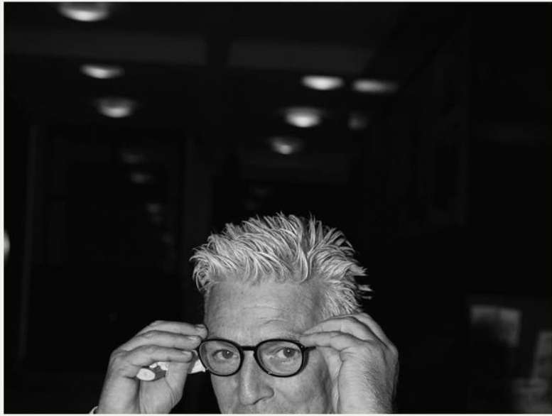 Black and white portrait of an adult with glasses adjusting them with his fingers, under bright recessed lighting indoors.