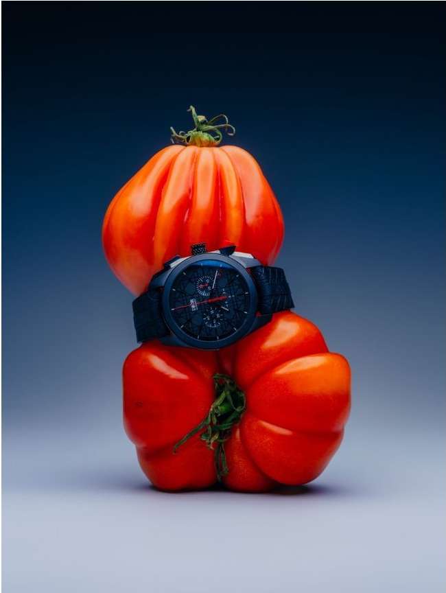 Two large heirloom tomatoes stacked vertically, with a blue-black wristwatch with a black band placed between them in a studio setting.