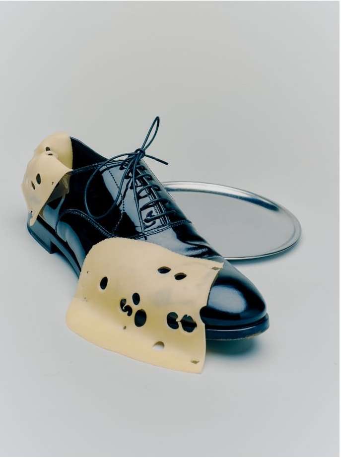 Black leather dress shoe sits next to sliced cheese with holes and a round, silver tray, on a white background in a studio shot.