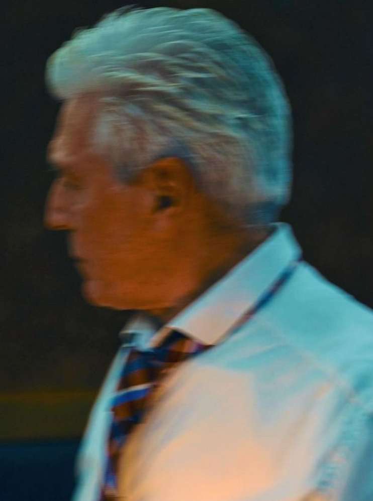 Older man with gray hair, wearing a white collared shirt and patterned tie, in a side-view, medium shot.