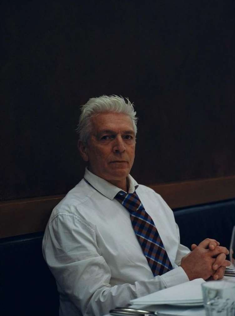 Portrait of a mature Caucasian man with gray hair wearing a shirt and tie, sitting at a table against a dark background.