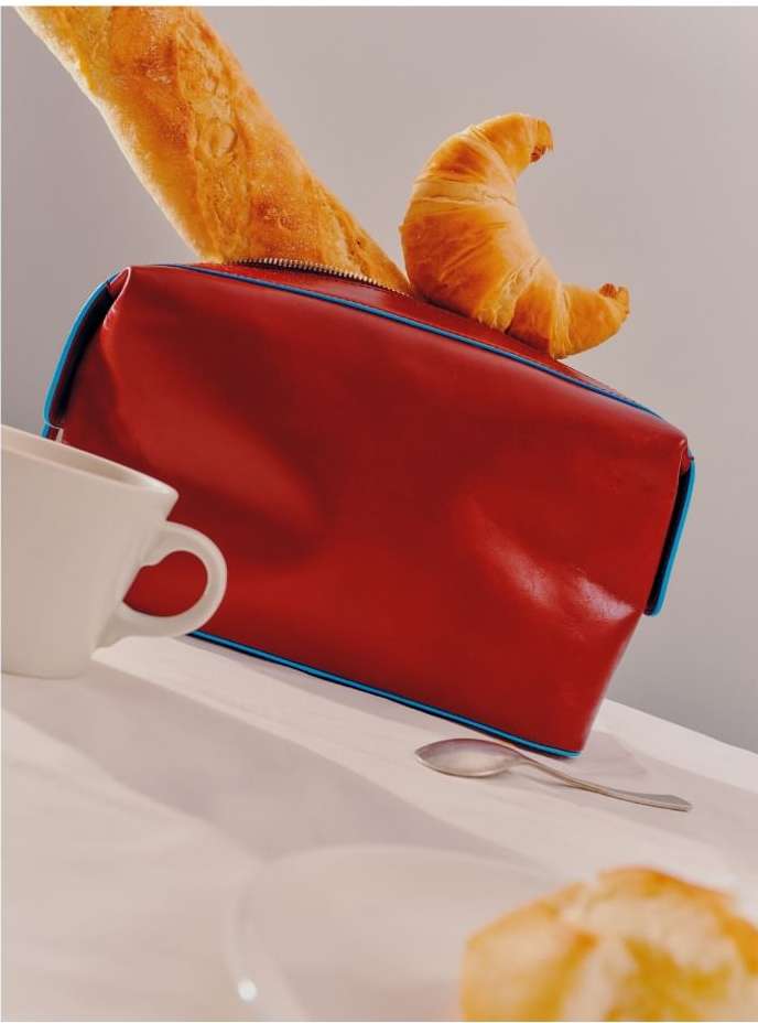 Red toiletry bag with a croissant and baguette on top, set on a white table with a cup, plate, and spoon. Food and travel concept.