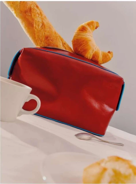 Red toiletry bag with a croissant and baguette on top, set on a white table with a cup, plate, and spoon. Food and travel concept.