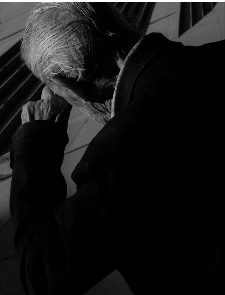 Monochrome portrait of an elderly person in shadow. Face partially visible with grey hair, hand near face and dark clothing.