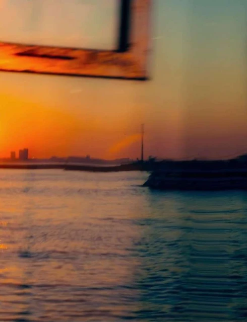Scenic view of water and cityscape at sunset, with orange sky, silhouetted buildings, and window frame detail.