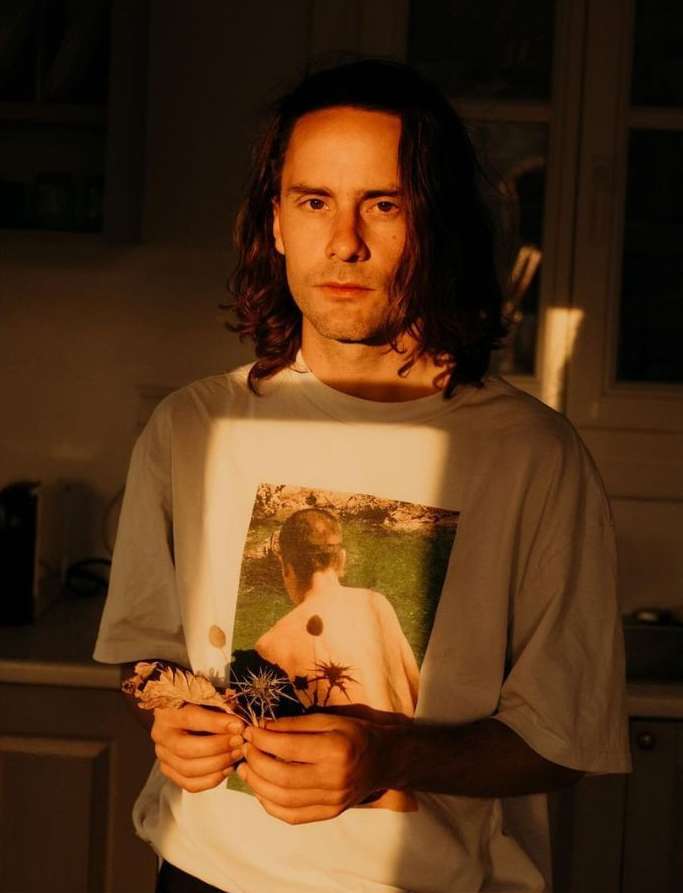 Portrait of a young adult male in a white t-shirt with a graphic print holding dried flowers in an indoor setting with golden sunlight.