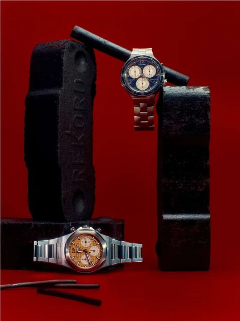 Two stainless steel watches on black stands and rods against a red backdrop create an industrial, stylized horology display.