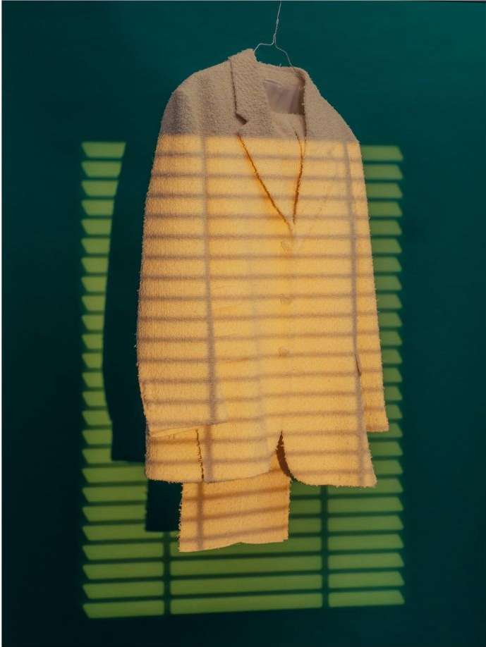 Beige suit hanging on wire hanger against green wall, featuring patterns of light and shadow from window blinds.