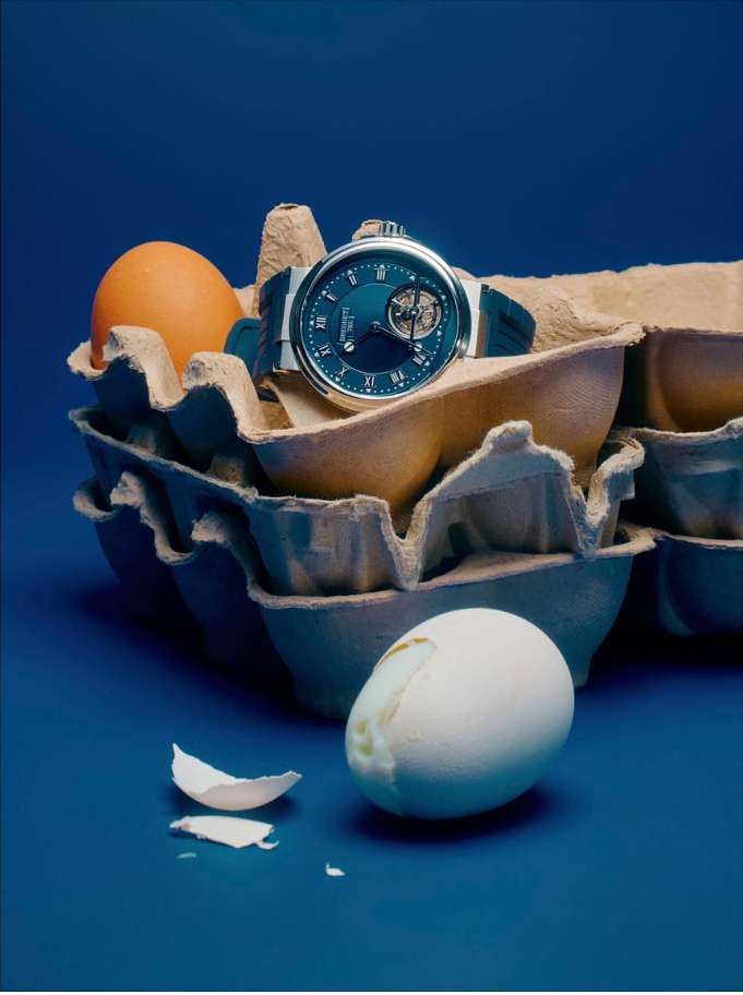 Luxury watch resting on an egg carton surrounded by raw eggs against a dark blue background in a studio lighting setting.