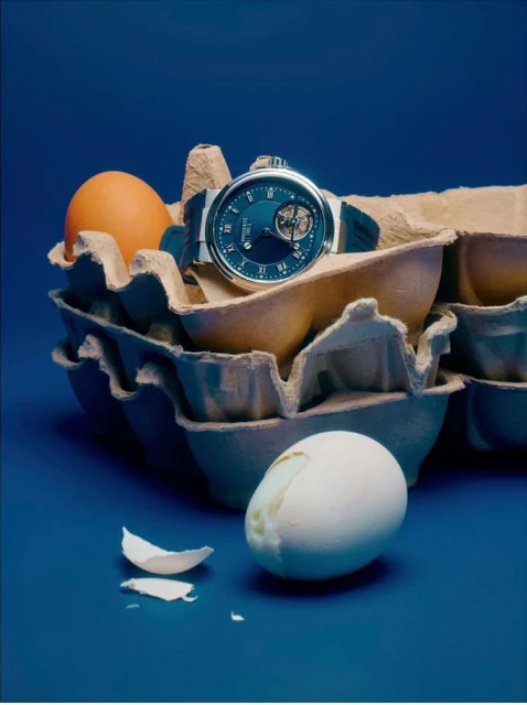 Luxury watch resting on an egg carton surrounded by raw eggs against a dark blue background in a studio lighting setting.