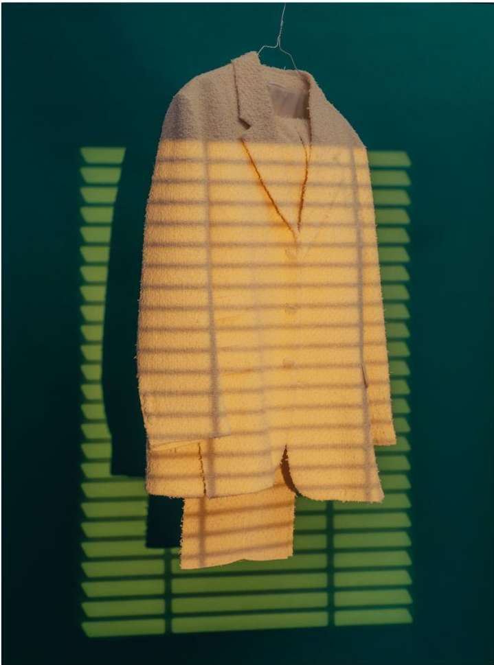 Suit hanging on a hanger against a green wall with sunlight and shadows casting horizontal lines creating a unique visual effect.