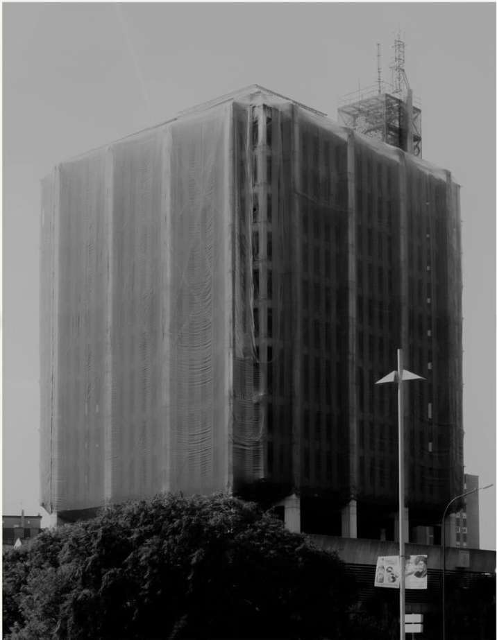 Tall building under construction, encased in scaffolding and protective netting. Urban scene with trees, photographed in black and white.