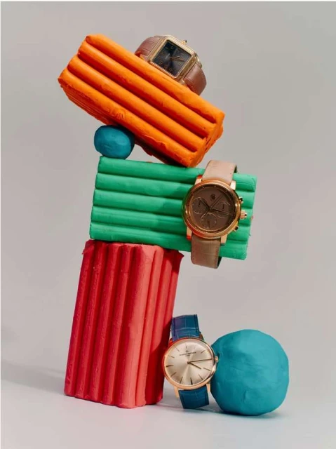 Still life composition of four wristwatches balanced on stacked colorful clay blocks against a light grey backdrop in a studio setting.
